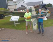 Two men holding the large-scale model of Major Helmut Wick’s Messerschmitt Bf 109E-4 in front of the Museum’s Spitfire