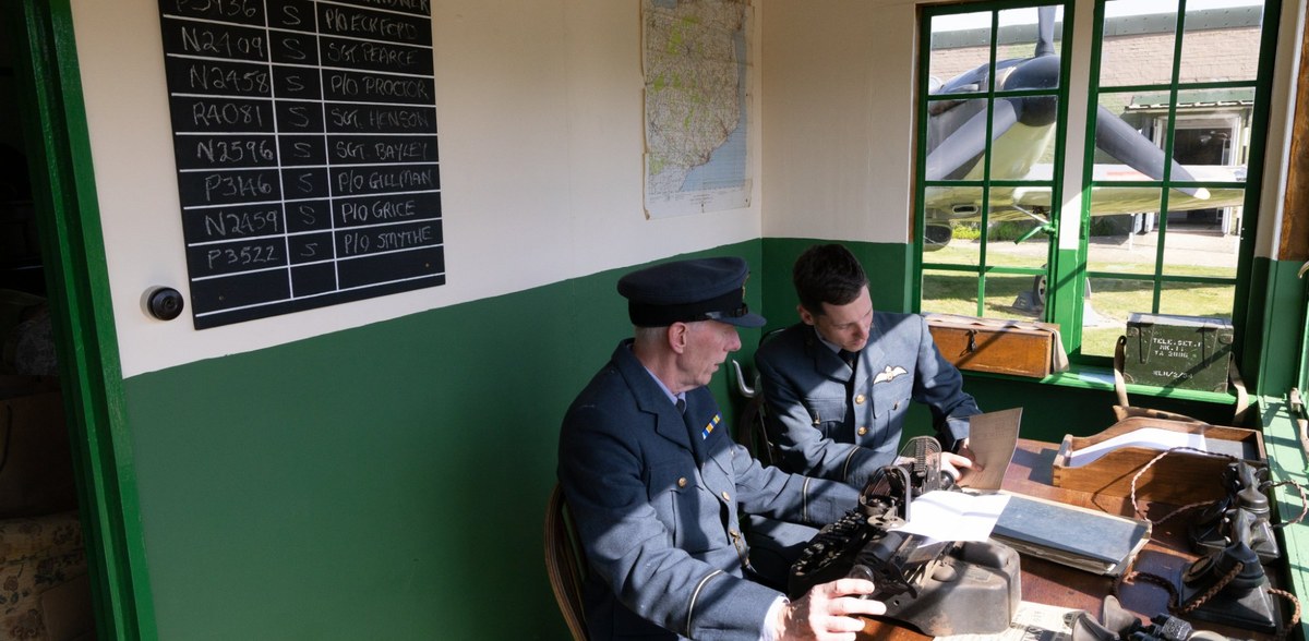 Volunteers in RAF uniform re-enacting the Operations Room, with a Spitfire visible through the window.