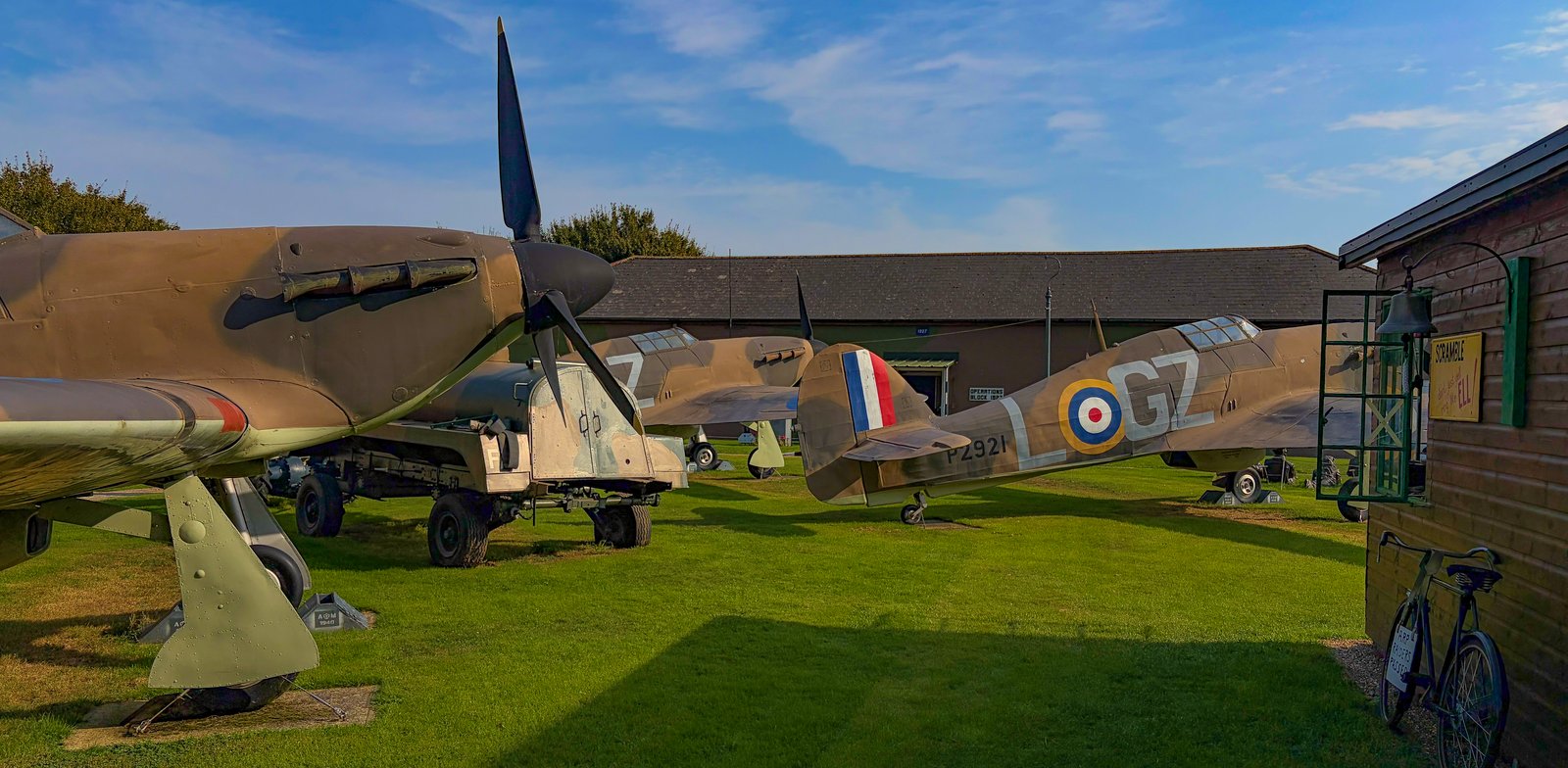 Replica Hurricanes and a Spitfire on the grass in the museum grounds, with a wartime bicycle and scramble bell by a hut