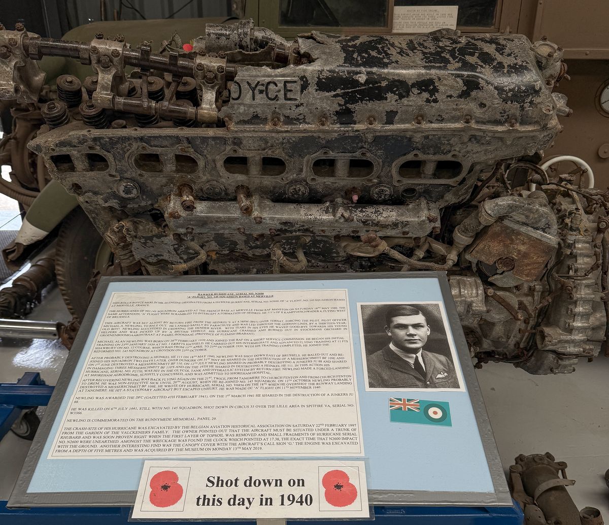A battered, excavated Rolls-Royce Merlin engine on display, with a panel telling the story of P/O Michael Newling, his photograph, an RAF flag and a 'Shot down on this day in 1940' sign with poppies