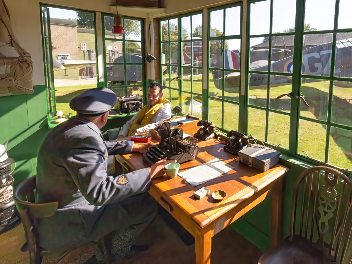 Two volunteers in RAF uniform at the Watch Office desk with a typewriter and field telephones, a Hurricane visible through the green-framed windows