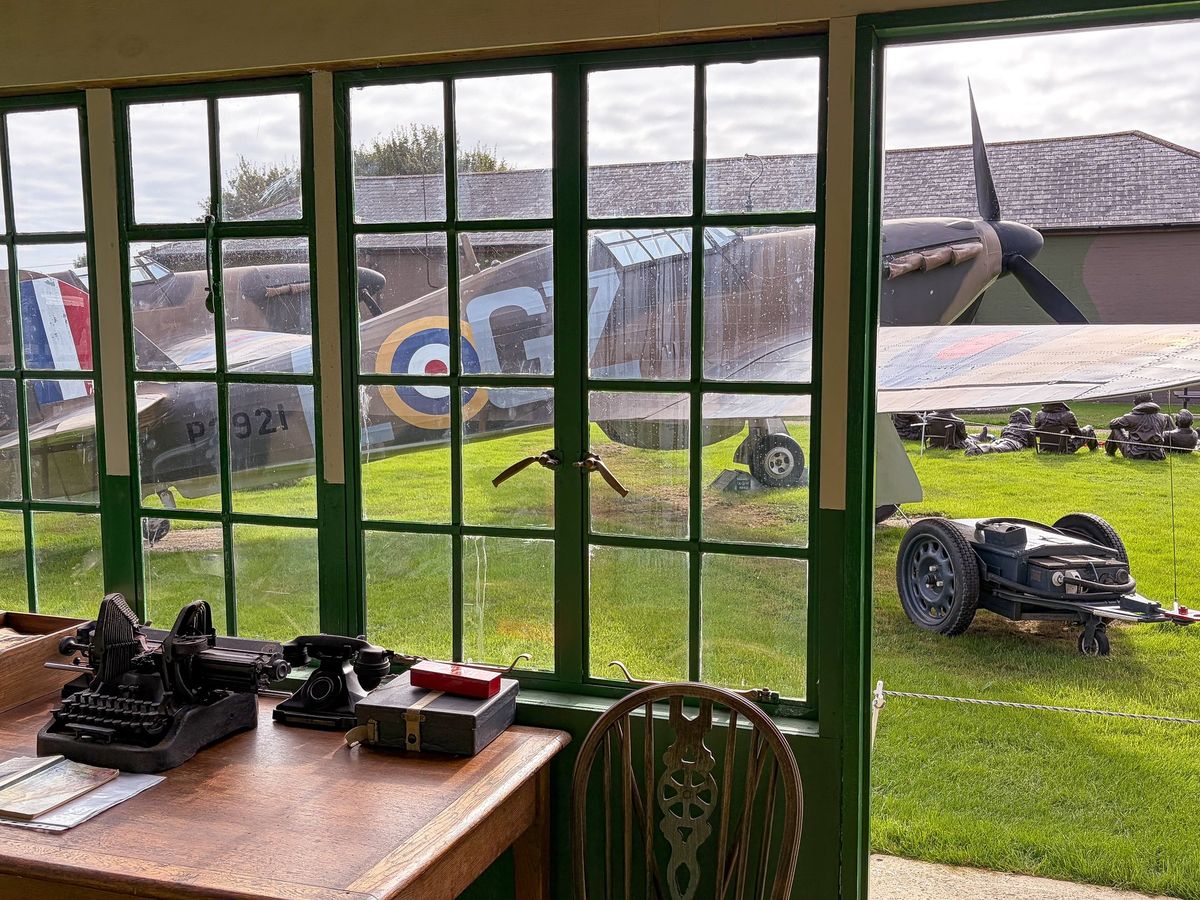 The Watch Office desk with field telephones and a typewriter, looking out through green-framed windows at a Hurricane in the museum grounds